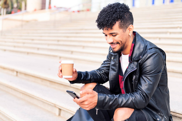 happy young latin student sitting on the stairs smiles consulting his phone with a coffee in his hand, concept of technology and urban lifestyle - Fotoğraf, Görsel