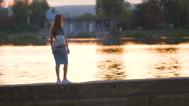 Achteraanzicht van eenzame jonge vrouw die alleen aan de oever van het meer geniet van warme avond. Welzijn en ontspanning in de natuur concept - Video