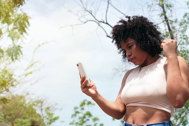 Женщина с афроволосами в парке пользуется телефоном. - Фото, изображение