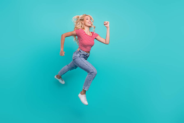 Photo of adorable shiny young lady wear striped t-shirt jumping high running fast looking empty space smiling isolated turquoise color background. - Zdjęcie, obraz