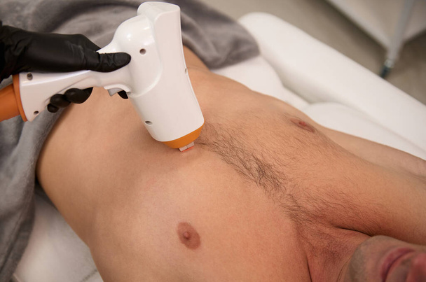 Overhead view of the torso of an athlete man during a hair removal procedure in a laser epilation studio. Laser hair removal for men using a modern ultrasonic hair removal machine. Male waxing - Fotó, kép