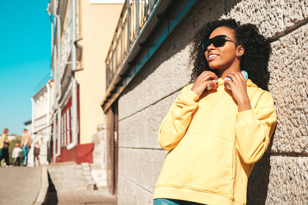 Beautiful black woman with afro curls hairstyle.Smiling model in yellow hoodie.Sexy carefree female enjoying listening music in wireless headphones.Posing on street background near wall in sunglasses - Foto, Imagen