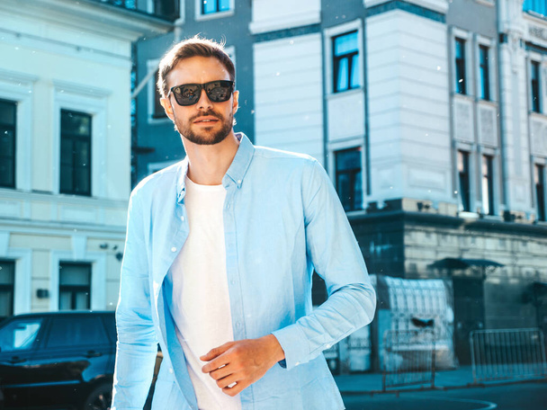 Handsome confident stylish hipster lambersexual model.Modern man dressed in blue shirt. Fashion male posing near skyscraper on the street background in sunglasses. Outdoors at sunset - Zdjęcie, obraz