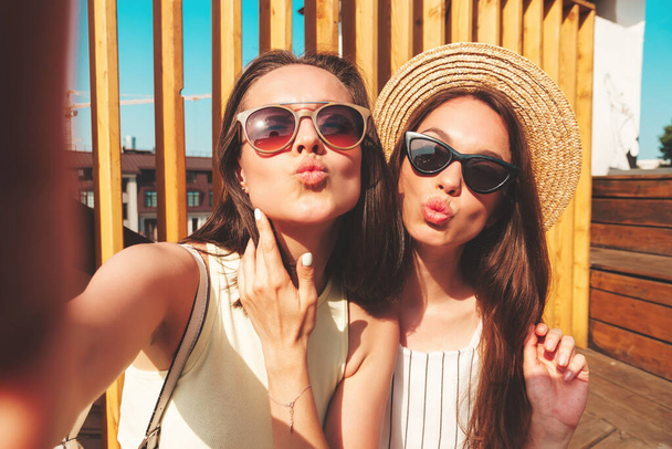 Two young beautiful smiling hipster female in trendy summer clothes.Sexy carefree women posing on the street background in hat. Positive pure models having fun at sunset, taking Pov selfie - Photo, Image