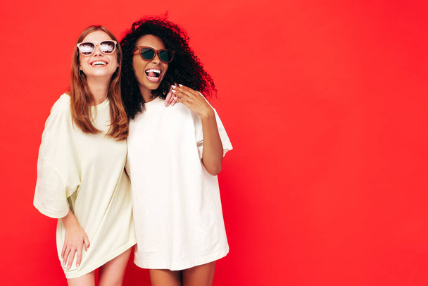 Dois jovens bonita sorridente internacional hipster fêmea em roupas de verão na moda. Mulheres despreocupadas Sexy posando perto da parede vermelha no estúdio. Modelos positivos se divertindo. Conceito de amizade. Em óculos escuros - Foto, Imagem