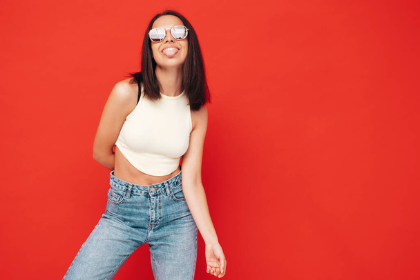 Portrait of young beautiful smiling female in trendy summer clothes. Sexy carefree woman posing near red wall in studio. Positive model having fun indoors. In sunglasses. Shows tongue - Foto, Imagem