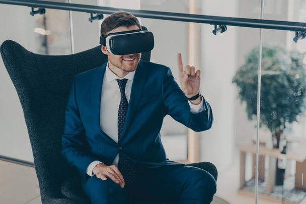 Εκπληκτικός επιχειρηματίας με επίσημο κοστούμι που κάθεται στο λόμπι της εταιρείας φορώντας γυαλιά εικονικής πραγματικότητας με ακουστικά - Φωτογραφία, εικόνα