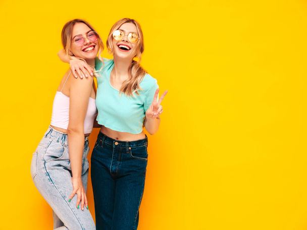 Two young beautiful smiling blond hipster female in trendy summer clothes. Sexy carefree women posing near yellow wall in studio. Positive models having fun. Cheerful and happy. In sunglasses - Foto, Bild