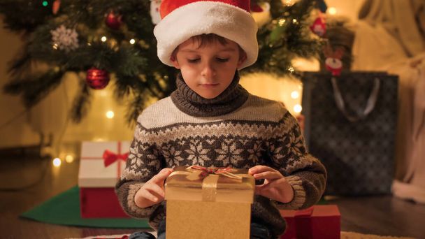 Aranyos fiú télapó kalapban, aki a karácsonyi ajándékdobozba néz. Karácsonyi varázslat. A téli ünnepeket ünneplő családok és gyermekek. - Fotó, kép