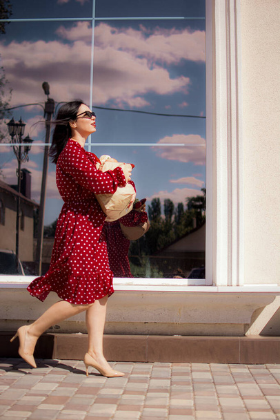 Happy grocery shopper. Portrait of beautiful traditional white woman in red dress holding paper shopping bag full of groceries. - Φωτογραφία, εικόνα