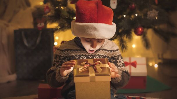 Тонированный портрет изумленного мальчика, смотрящего в светящуюся коробку с подарками на Рождество ночью - Фото, изображение