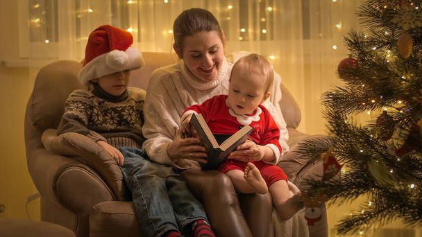 Gelukkig lachende moeder met kinderen die sprookjesboek lezen in fauteuil naast kerstboom in de woonkamer. Gezinnen en kinderen vieren wintervakantie. - Foto, afbeelding