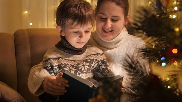 Happy smiling boy with mother reading fairy tale book next to glowing decorated Christmas tree. Families and children celebrating winter holidays. - Foto, Imagen