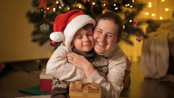 幸せな笑顔の母と息子の肖像サンタの帽子抱擁クリスマスツリーの隣に家 - 写真・画像