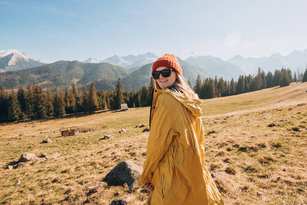 Молодая блондинка в солнечных очках танцует в горах. Свобода, счастье, путешествия и отпуск, активный отдых. Счастливая девушка в красной шляпе и желтой куртке - Фото, изображение