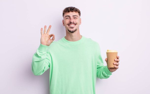 young handsome man feeling happy, showing approval with okay gesture. take away coffee concept - Photo, image