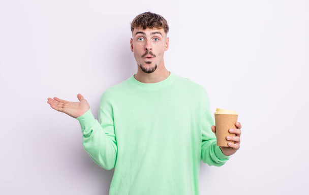 young handsome man looking surprised and shocked, with jaw dropped holding an object. take away coffee concept - Фото, зображення