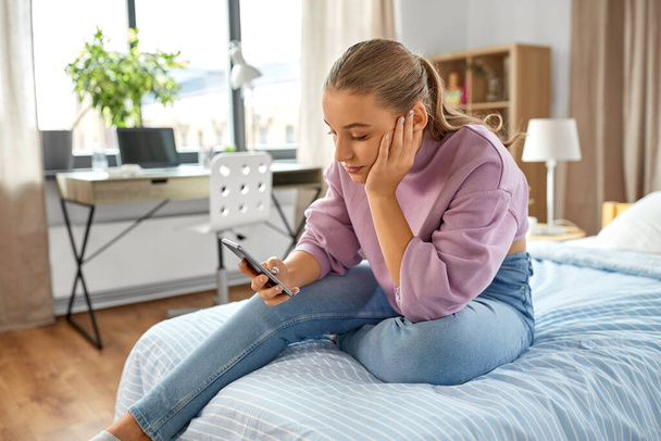 sad girl with smartphone sitting on bed at home - Фото, изображение