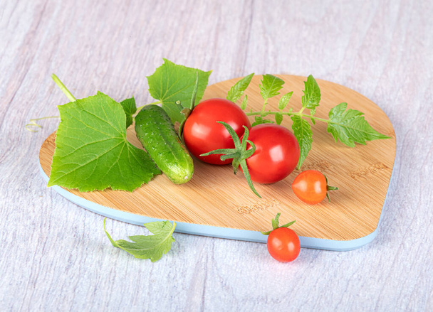 Ingredientes de ensalada (tomates cherry, pepinos, pepinillos, verduras) en una tabla de cortar de madera sobre un fondo claro - Foto, Imagen