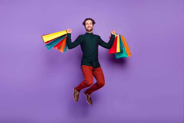 Photo of impressed lucky young guy dressed smart casual clothes jumping high holding shopping bags isolated violet color background. - Zdjęcie, obraz
