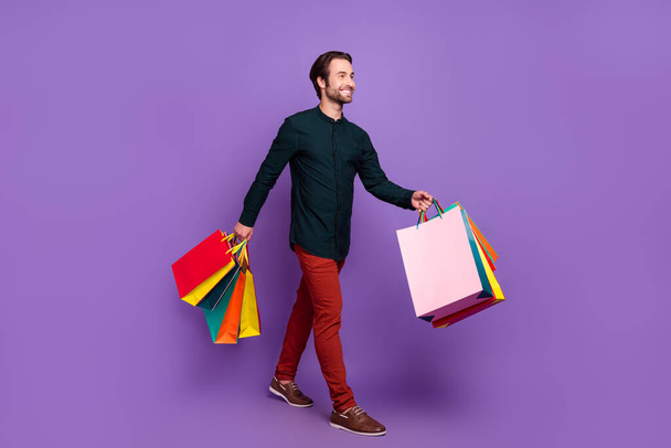 Photo of pretty charming young guy dressed smart casual clothes walking holding shopping bargains smiling isolated violet color background. - Foto, afbeelding