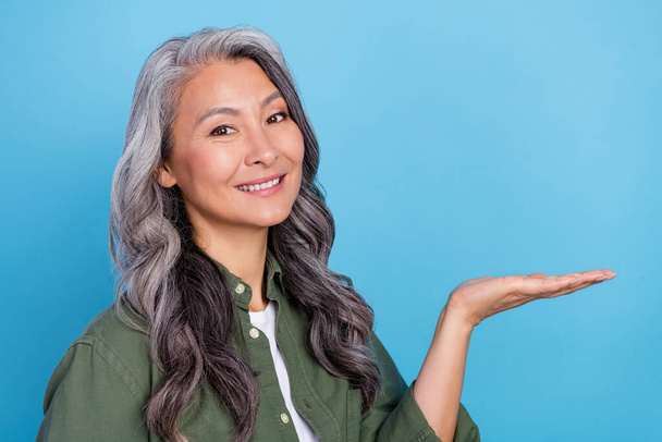 Photo of cute elder grey hairdo lady hold empty space wear green shirt isolated on blue color background. - Foto, imagen