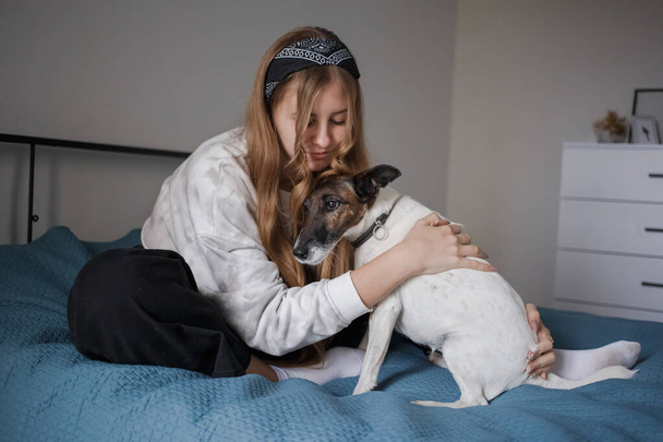 A young 20s girl with a headband and long blond hair sits on a bed in her living room and hugs a small white dog - 写真・画像