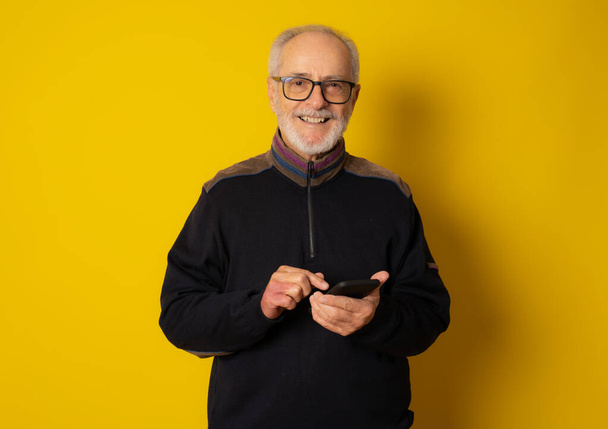 Концепция образа жизни, технологий и пожилых людей: красивый старый седой мужчина, одетый в элегантный повседневный стиль с помощью смартфона и улыбающийся на желтом фоне - Фото, изображение
