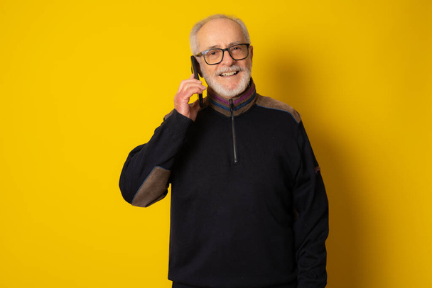 Yaşam tarzı, teknoloji ve yaşlılar konsepti: Yakışıklı, gri saçlı, şık giyinmiş, akıllı telefon kullanan ve sarı arka planda gülümseyen adam. - Fotoğraf, Görsel