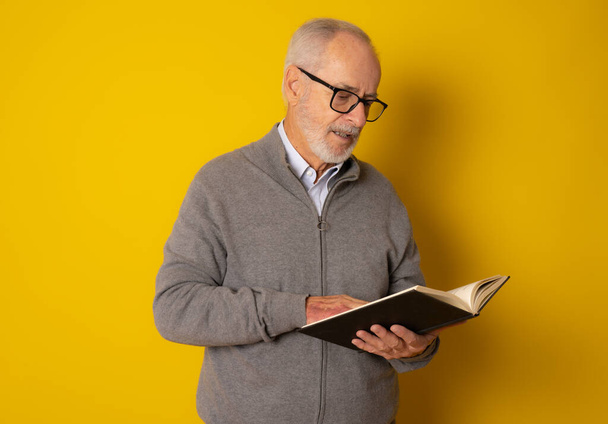 Mature smiling man reading a book isolated over yellow background. - 写真・画像