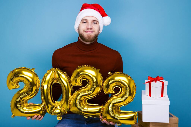 Ein Mann mit rotem Hut hält die Zahlen für das kommende Jahr 2022 in den Händen. Kisten mit Geschenken liegen in der Nähe - Foto, Bild