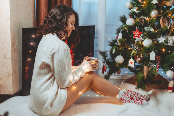 young pretty woman sitting near christmas tree drinking cup of coffee copy space - Foto, Bild