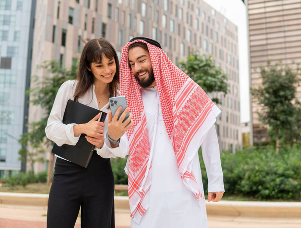 Усміхнені різні ділові люди арабський чоловік і біла жінка, використовуючи смартфон на відкритому повітрі
 - Фото, зображення