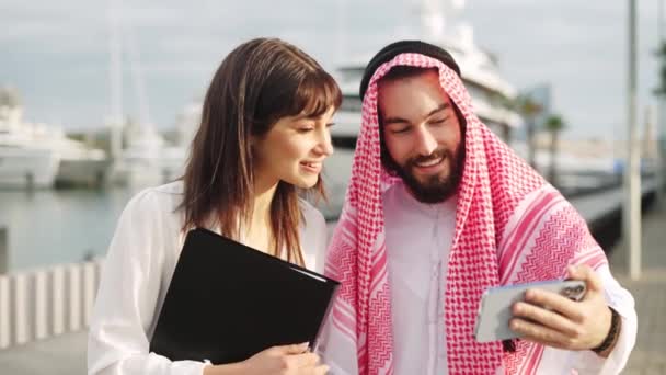 Mosolygó arab üzletember mutat valamit az okostelefonon vonzó európai női asszisztensének - Felvétel, videó