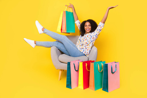 Full body photo of sweet young wavy hairdo lady sit on armchair with bags wear shirt jeans sneakers isolated on yellow background - Фото, изображение