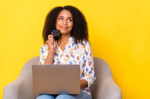 Photo of cute millennial curly hairdo lady hold laptop card look empty space sit on armchair wear shirt jeans isolated on yellow background - Foto, immagini