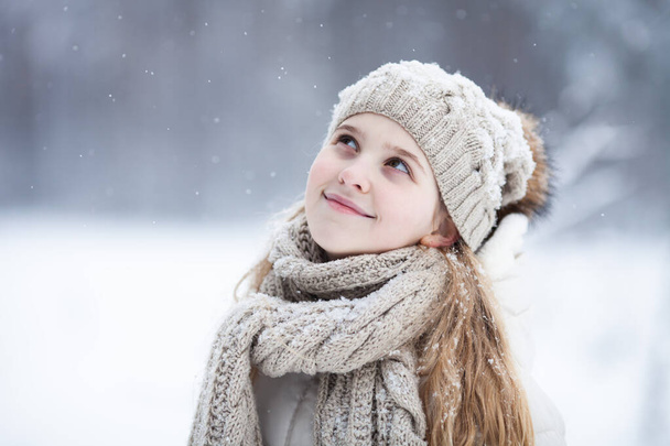 Πορτρέτο ενός χαριτωμένο ξανθό κορίτσι φορώντας ζεστό πλεκτό καπέλο και μαντήλι κοιτάζοντας προς τα πάνω στο χειμερινό χιονισμένο φόντο. Αγάπη χειμώνας, κάνε μια ευχή, τα όνειρα γίνονται πραγματικότητα. - Φωτογραφία, εικόνα