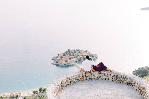 Kotor, Montenegro - 08.06.17: Bräutigam im weißen Hemd und Braut im weinroten Kleid sitzen auf der Aussichtsplattform und blicken auf die Insel Sveti Stefan hinunter. Budva, Montenegro - Foto, Bild