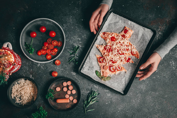 Children hand holds a baking dish with a Christmas tree pizza on a dark background with cherry tomatoes, sausages and mozzarella cheese, close-up side view. The concept of preparing for Christmas and New Year. - Photo, Image