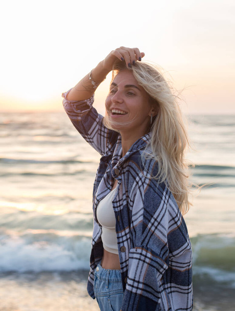 bela menina cabelo loiro feliz em uma camisola está de pé descalço na praia ao nascer do sol luz  - Foto, Imagem