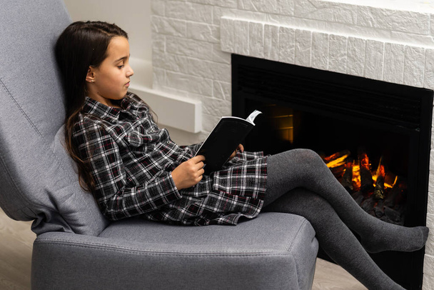 Little girl in dress sits in chair with book near fireplace. - Photo, Image