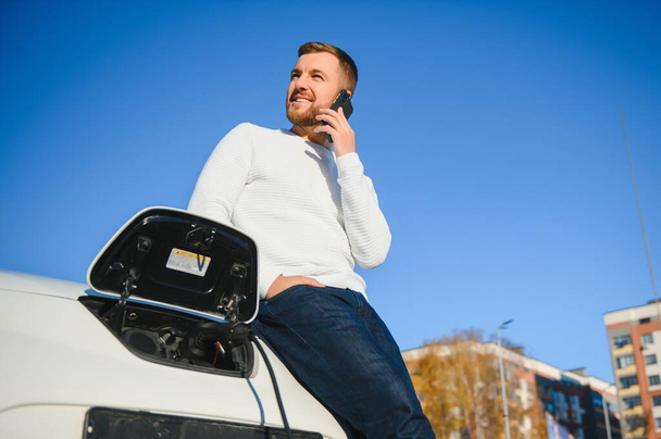 The guy sat down on the hood of the car. His car is charging at the charging station. A man looks at the smartphone screen and smiles. - Photo, image
