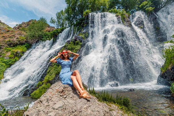 A happy female traveler is resting near the beautiful and large Shaki waterfall in Armenia. Fresh water and tourist attractions concept - Photo, Image