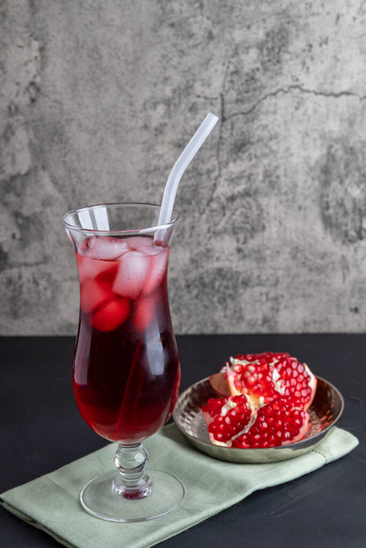 Een hoog glas rood sap met ijs en rietjes op een grijze achtergrond, naast een rijpe granaatappelvrucht. Verticale positionering, kopieerruimte - Foto, afbeelding