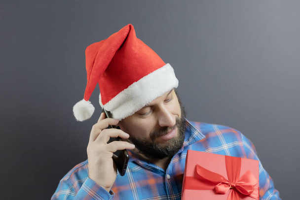 A man in a santa hat with a red box with a gift talking on a cell phone - Zdjęcie, obraz