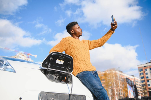 Portrait of young handsome black man in business suit, making selfie photo on his smartphone while leaning on his electric car, charging the battery at city power station - 写真・画像