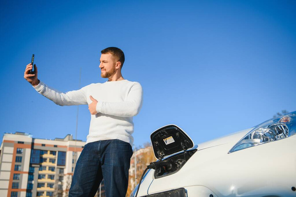 Portrait of young handsome european man, making selfie photo on his smartphone while leaning on his electric car, charging the battery at city power station after buying frsh products in trade mall - Foto, Imagen