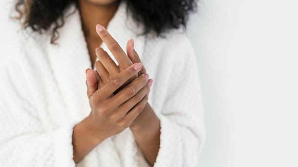 Cropped view of young afro american woman in bathrobe applying hand cream, standing against white copy space background. Soft skin, skincare, spa and manicure concept - Photo, Image
