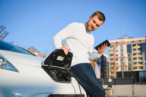 young handsome man holding charging cable at electric charging station point standing near his new car. - Valokuva, kuva