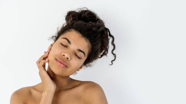 Beauty face. Web banner, portrait of young african american woman with natural makeup touch healthy, fresh, glowing and hydrated skin. Female stand with naked shoulder on white background, copy space - 写真・画像